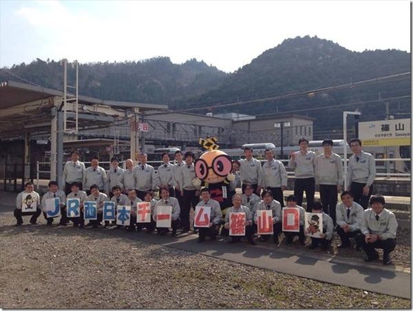 JR篠山口駅の職員が一文字ずつ入ったパネルを持って踏切のゆるキャラと一緒に駅のホームの前で記念撮影している写真